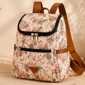 May include: A backpack featuring a cream-colored base with a dense floral pattern of pink, red, and green roses. It includes brown adjustable shoulder straps, black zipper closures, a front zippered pocket, and a triangular brown patch at the base.
