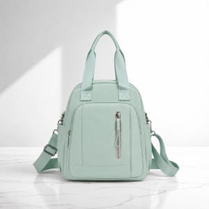 May include: A mint green backpack with two top handles and a vertical zipped front pocket. The bag features adjustable side straps with metal hardware, a main zipped compartment, and a smooth, solid-coloured exterior finish throughout.