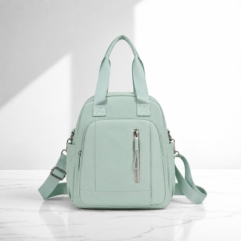 May include: A mint green backpack with two top handles and a vertical zipped front pocket. The bag features adjustable side straps with metal hardware, a main zipped compartment, and a smooth, solid-coloured exterior finish throughout.