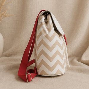 May include: A bucket-style backpack featuring a beige and white chevron pattern. The bag includes a solid white top flap, a drawstring closure, and bright red adjustable fabric shoulder straps with silver-toned metal hardware buckles.