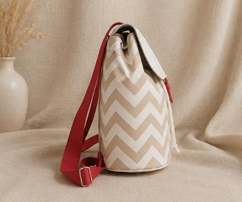 May include: A bucket-style backpack featuring a beige and white chevron pattern. The bag includes a solid white top flap, a drawstring closure, and bright red adjustable fabric shoulder straps with silver-toned metal hardware buckles.