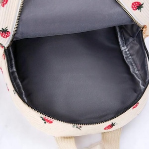 May include: A white corduroy backpack with a strawberry print. The backpack has a zipper closure and a gray lining.