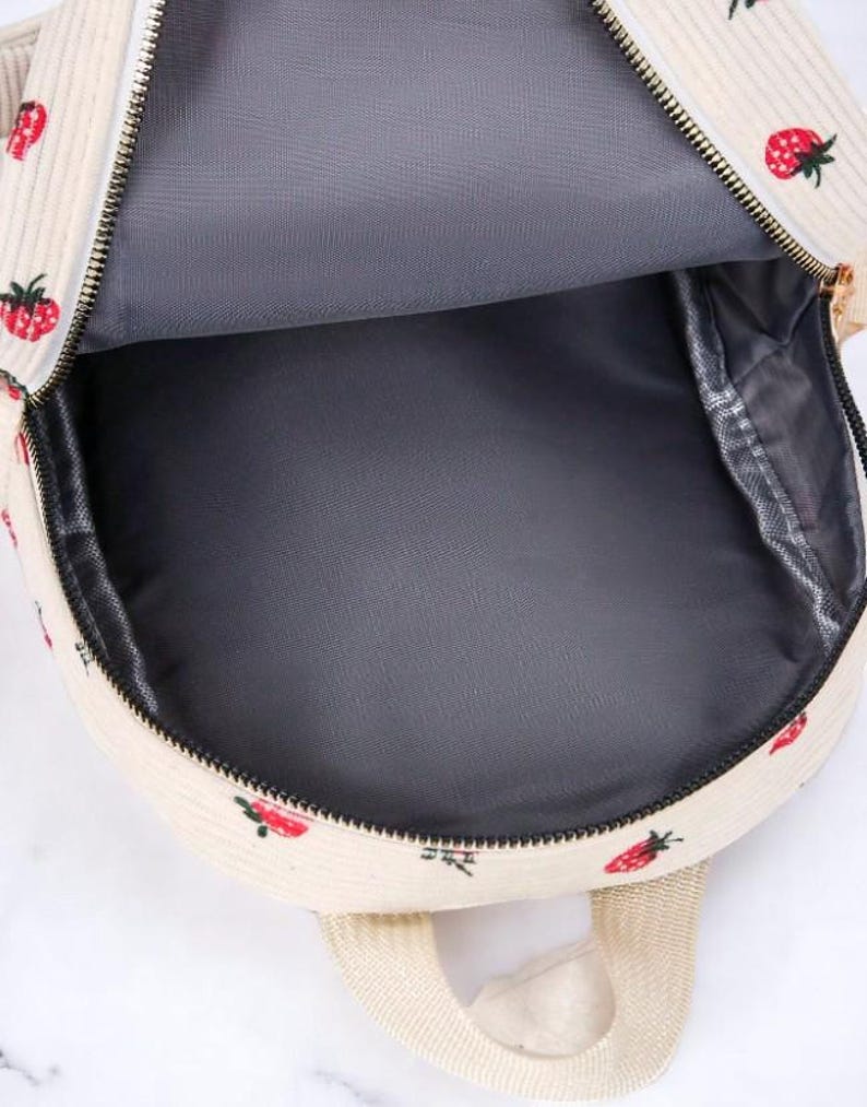 May include: A white corduroy backpack with a strawberry print. The backpack has a zipper closure and a gray lining.