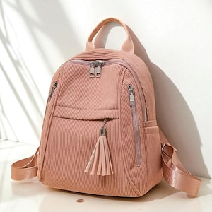 May include: A pink corduroy backpack with silver zippers and a tassel. The backpack has a top handle, adjustable straps, and multiple zippered compartments. The bag is approximately 12 inches tall and 10 inches wide.