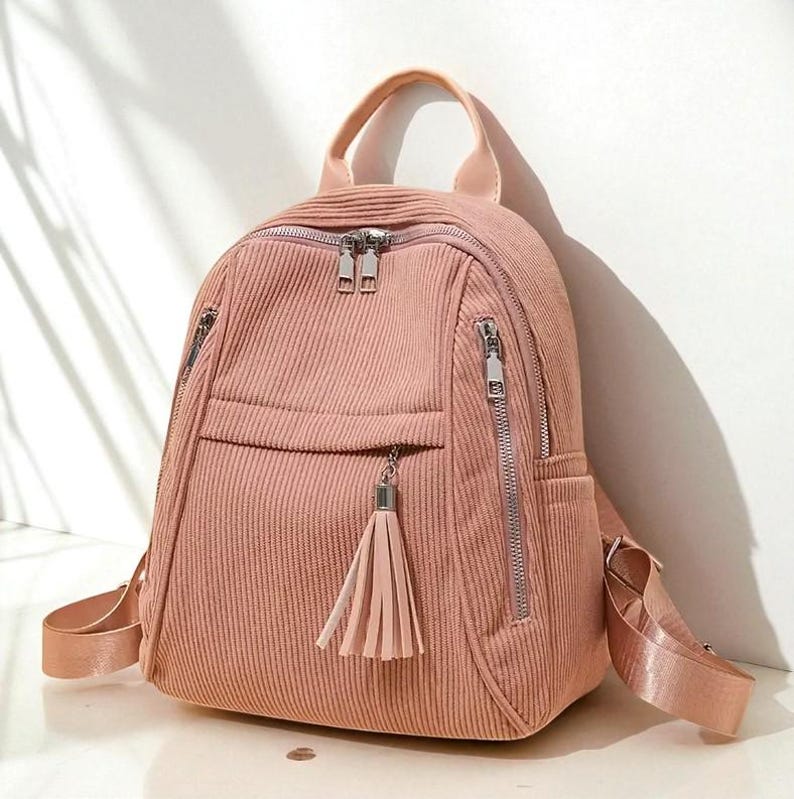 May include: A pink corduroy backpack with silver zippers and a tassel. The backpack has a top handle, adjustable straps, and multiple zippered compartments. The bag is approximately 12 inches tall and 10 inches wide.
