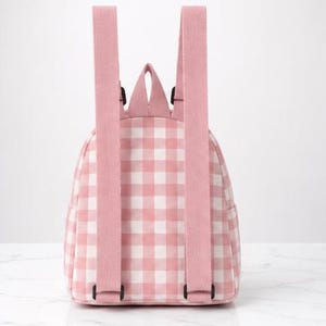 May include: A small backpack featuring a light pink and white gingham check pattern. The bag includes two adjustable pink shoulder straps, a top carry handle, and a main compartment with a zip closure, shown from the back view.