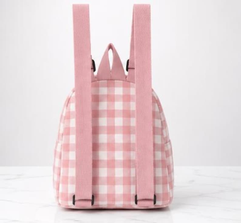 May include: A small backpack featuring a light pink and white gingham check pattern. The bag includes two adjustable pink shoulder straps, a top carry handle, and a main compartment with a zip closure, shown from the back view.