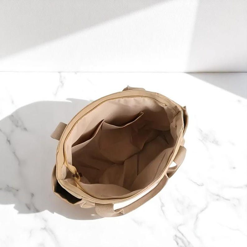 May include: Beige canvas tote bag with a zippered top and interior pockets. The bag has sturdy handles and a neutral color scheme, suitable for everyday use. The interior is a lighter shade of beige.