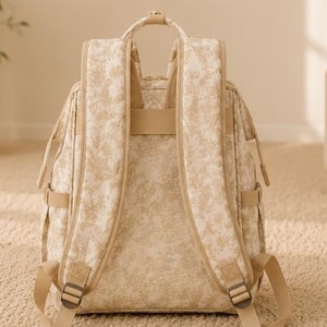 Peut inclure: Un sac &agrave; dos beige et cr&egrave;me arborant un motif floral d&eacute;licat sur toute sa surface ext&eacute;rieure. Le sac comprend deux bretelles rembourr&eacute;es, une poign&eacute;e de transport sup&eacute;rieure et des sangles r&eacute;glables avec des boucles en m&eacute;tal.