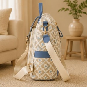 May include: A side view of a backpack featuring a beige and white geometric diamond pattern with light blue accents. The bag includes a top handle, blue side pockets, gold-toned metal hardware, and adjustable cream-colored fabric shoulder straps.