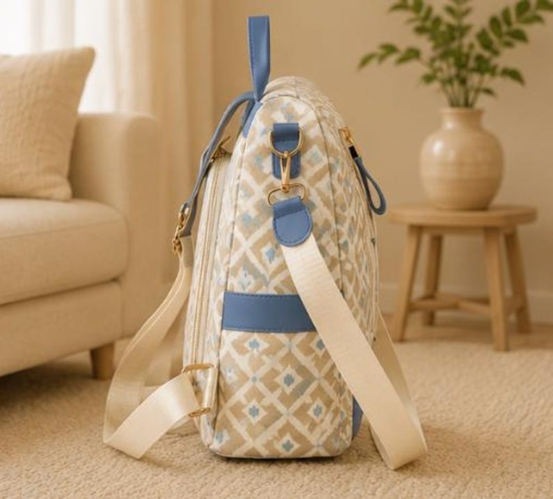 May include: A side view of a backpack featuring a beige and white geometric diamond pattern with light blue accents. The bag includes a top handle, blue side pockets, gold-toned metal hardware, and adjustable cream-colored fabric shoulder straps.