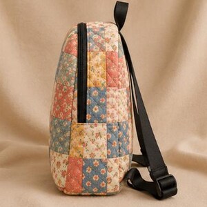 K&ouml;nnte beinhalten: Ein gesteppter Rucksack mit einem Patchwork-Design aus Blumenmustern in Blau-, Gelb- und Korallent&ouml;nen. Die Tasche verf&uuml;gt &uuml;ber einen schwarzen Rei&szlig;verschluss an der Seite, einen schwarzen Tragegriff und verstellbare schwarze Nylongurte.