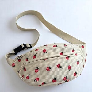 May include: Beige corduroy fanny pack with a repeating pattern of red strawberries. The bag has a beige adjustable strap, a black buckle, and a zippered front pocket. The fanny pack is designed to be worn around the waist or across the body.