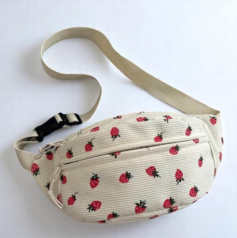 May include: Beige corduroy fanny pack with a repeating pattern of red strawberries. The bag has a beige adjustable strap, a black buckle, and a zippered front pocket. The fanny pack is designed to be worn around the waist or across the body.