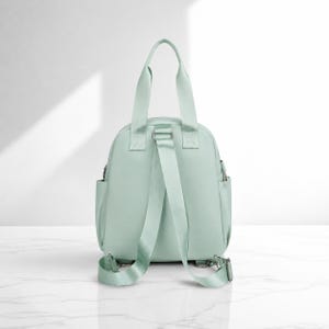 May include: A mint green backpack featuring two top carry handles and adjustable shoulder straps. The bag has a rounded main compartment with side pockets and silver-toned metal hardware attachments at the base of the straps.
