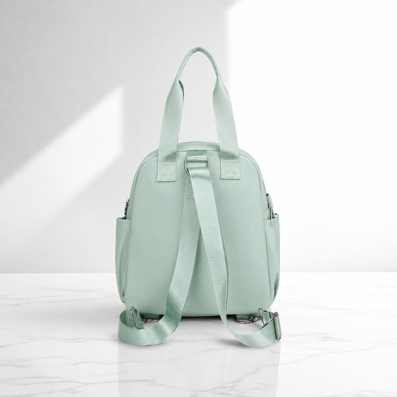 May include: A mint green backpack featuring two top carry handles and adjustable shoulder straps. The bag has a rounded main compartment with side pockets and silver-toned metal hardware attachments at the base of the straps.