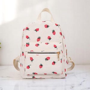 May include: A white corduroy backpack with a strawberry print. The backpack has a front zipper pocket and adjustable straps.
