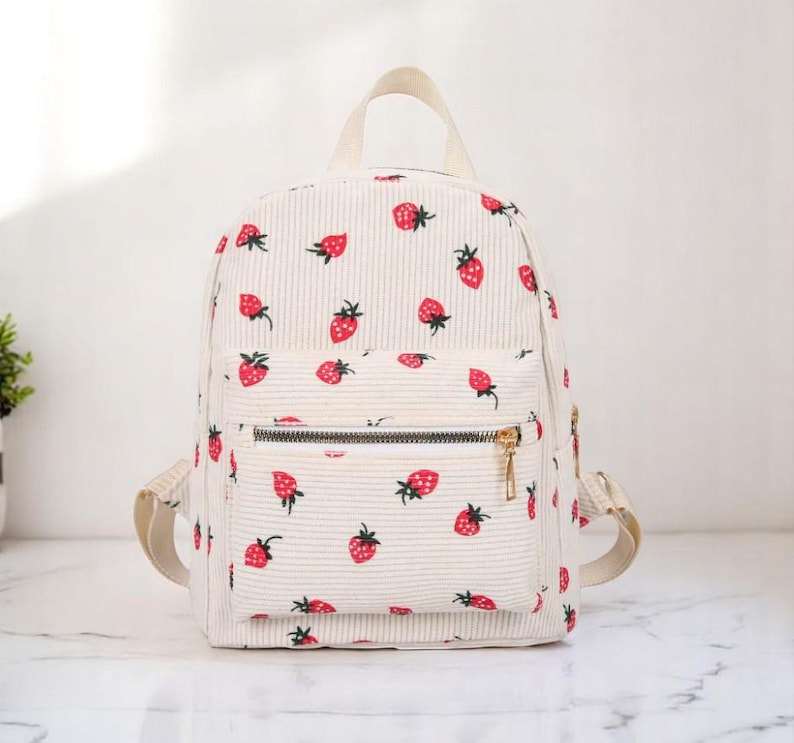 May include: A white corduroy backpack with a strawberry print. The backpack has a front zipper pocket and adjustable straps.