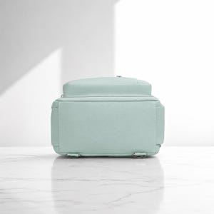 May include: A light mint green backpack shown from the back view. The bag features a rectangular main compartment with a flat base, visible stitching along the edges, and two small metal rings at the bottom corners for attaching shoulder straps.