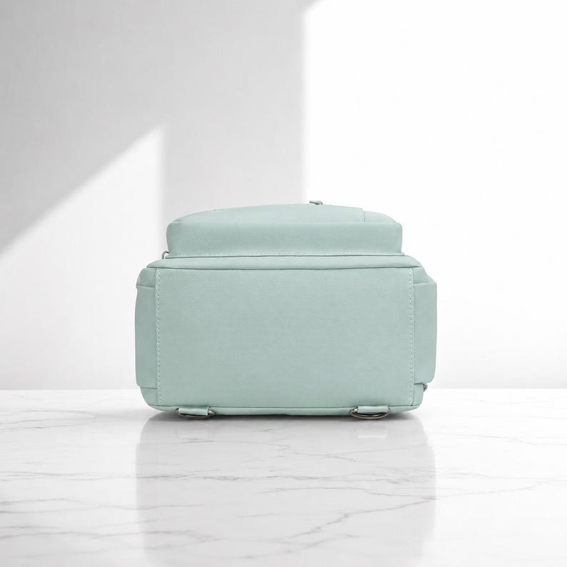 May include: A light mint green backpack shown from the back view. The bag features a rectangular main compartment with a flat base, visible stitching along the edges, and two small metal rings at the bottom corners for attaching shoulder straps.
