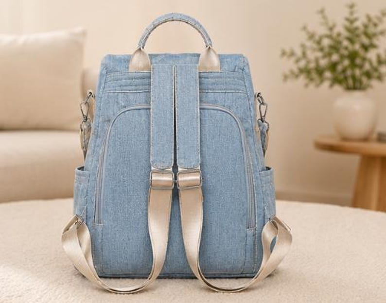 Puede incluir: Una mochila de mezclilla azul claro con un asa superior, dos correas beige ajustables y m&uacute;ltiples compartimentos con cremallera. La bolsa incluye bolsillos laterales y herrajes met&aacute;licos plateados, con un dise&ntilde;o estructurado y costuras visibles.