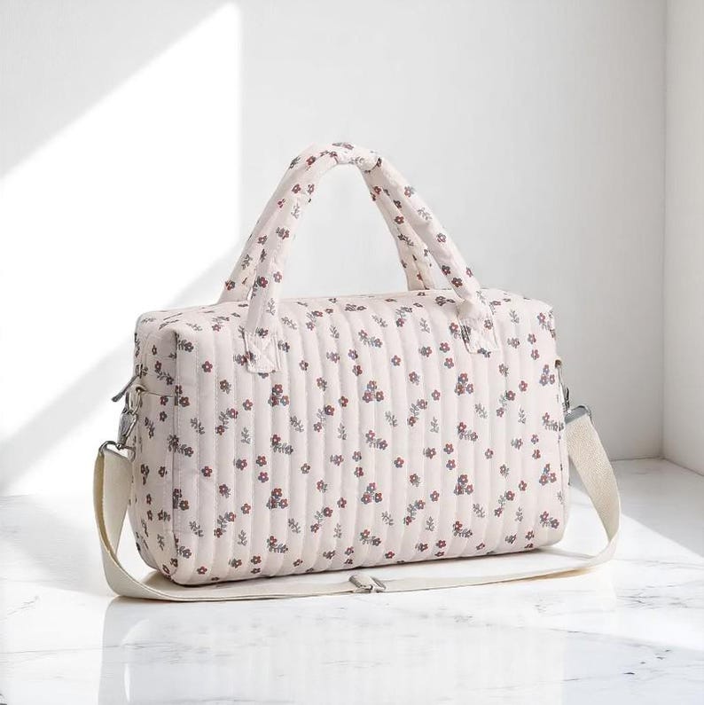 May include: A quilted duffel bag in a light cream color featuring a repeating pattern of small red and dark green floral sprigs. The bag has two padded top handles, a rectangular structure, and an adjustable off-white shoulder strap.