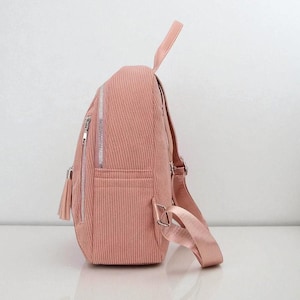 May include: A pink corduroy backpack with silver zippers and a tassel. The backpack has adjustable straps and a top handle. The bag has a front pocket and a main compartment. The corduroy fabric has a vertical ribbed texture.