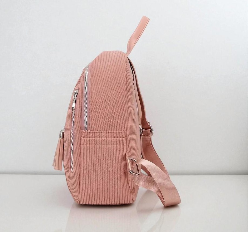 May include: A pink corduroy backpack with silver zippers and a tassel. The backpack has adjustable straps and a top handle. The bag has a front pocket and a main compartment. The corduroy fabric has a vertical ribbed texture.
