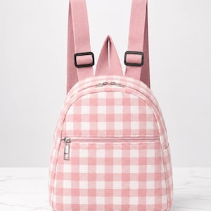 May include: A small backpack featuring a pink and white gingham chequered pattern. The bag includes two adjustable pink shoulder straps with black plastic buckles, a main zipped compartment, and a smaller front zipped pocket with a silver pull.
