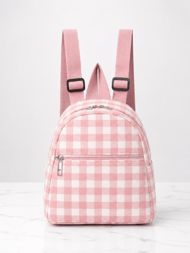 May include: A small backpack featuring a pink and white gingham chequered pattern. The bag includes two adjustable pink shoulder straps with black plastic buckles, a main zipped compartment, and a smaller front zipped pocket with a silver pull.