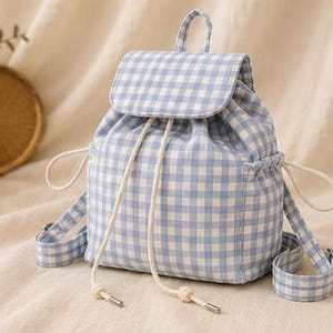 May include: A light blue and white gingham patterned backpack featuring a fold-over flap with a magnetic closure, a drawstring top with white cord ties, a top carry handle, and adjustable shoulder straps with metal hardware buckles.