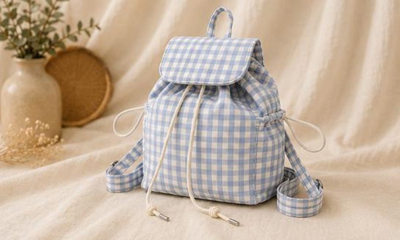 May include: A light blue and white gingham patterned backpack featuring a fold-over flap with a magnetic closure, a drawstring top with white cord ties, a top carry handle, and adjustable shoulder straps with metal hardware buckles.