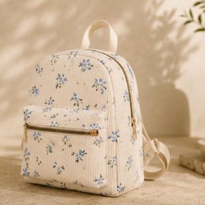 K&ouml;nnte beinhalten: Ein cremefarbener Mini-Rucksack aus Kord mit einem zarten blauen Blumenmuster. Die Tasche verf&uuml;gt &uuml;ber einen Tragegriff, verstellbare Schultergurte und eine Fronttasche mit Rei&szlig;verschluss sowie goldfarbene Beschl&auml;ge. Das Hauptfach hat einen Rei&szlig;verschluss.