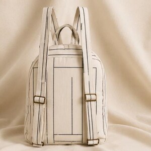 May include: A cream-colored backpack featuring a black line-drawn geometric pattern. The bag includes two adjustable shoulder straps with metallic buckles, a top handle, and a main compartment with a zipper closure, all made from fabric.