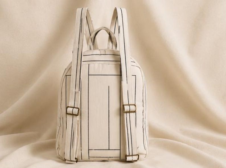 May include: A cream-colored backpack featuring a black line-drawn geometric pattern. The bag includes two adjustable shoulder straps with metallic buckles, a top handle, and a main compartment with a zipper closure, all made from fabric.
