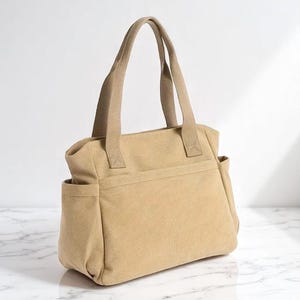May include: A tan canvas tote bag with two handles and two side pockets. The bag has a rectangular shape and a neutral color, suitable for various uses. The handles are a matching tan color.