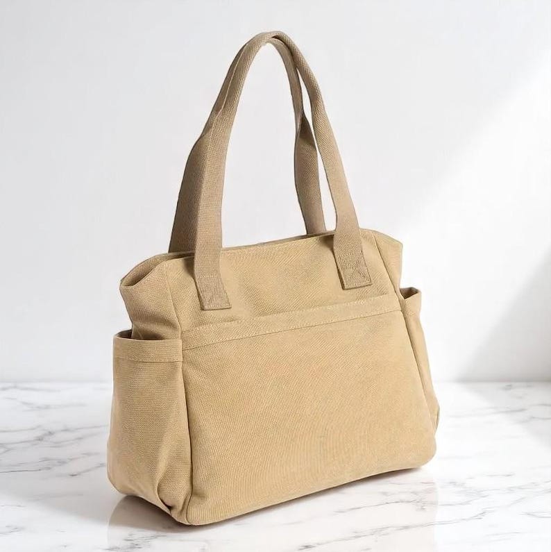 May include: A tan canvas tote bag with two handles and two side pockets. The bag has a rectangular shape and a neutral color, suitable for various uses. The handles are a matching tan color.