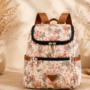May include: A backpack featuring a cream-colored base with a floral pattern of pink roses and green leaves. It includes brown shoulder straps, black zipper closures, a front zippered pocket, and a small triangular brown patch at the bottom center.