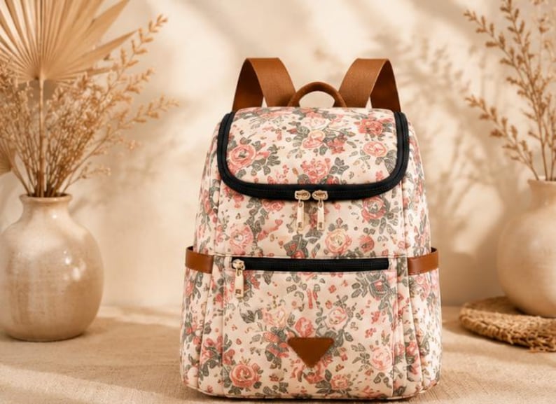 May include: A backpack featuring a cream-colored base with a floral pattern of pink roses and green leaves. It includes brown shoulder straps, black zipper closures, a front zippered pocket, and a small triangular brown patch at the bottom center.