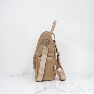 K&ouml;nnte beinhalten: Ein beigefarbener Rucksack aus Kordstoff in Seitenansicht mit vertikaler Rippenstruktur, einem Tragegriff, einer Seitentasche und verstellbaren, hellbeigen Gurtb&auml;ndern mit silberfarbenen Metallbeschl&auml;gen sowie einem geflochtenen Rei&szlig;verschlussanh&auml;nger.