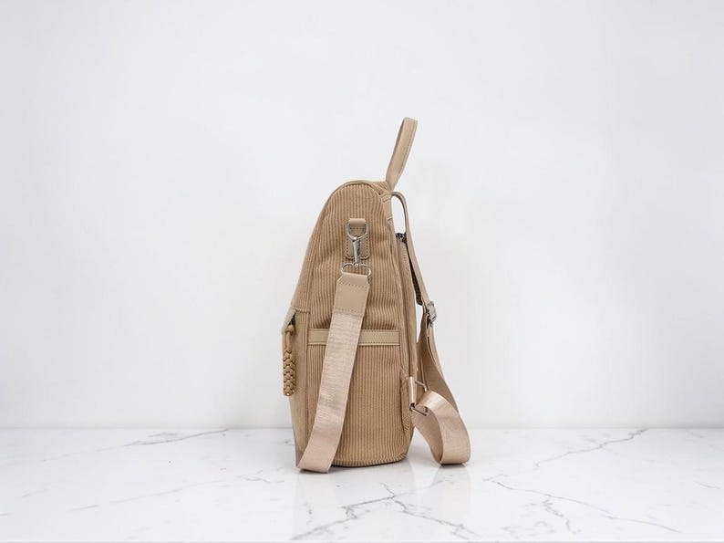 K&ouml;nnte beinhalten: Ein beigefarbener Rucksack aus Kordstoff in Seitenansicht mit vertikaler Rippenstruktur, einem Tragegriff, einer Seitentasche und verstellbaren, hellbeigen Gurtb&auml;ndern mit silberfarbenen Metallbeschl&auml;gen sowie einem geflochtenen Rei&szlig;verschlussanh&auml;nger.