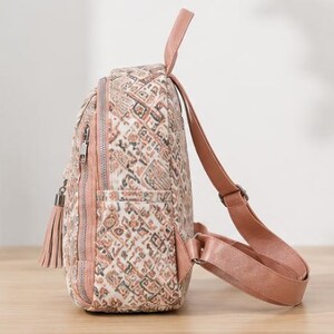May include: A small backpack featuring a multi-colored geometric pattern in shades of pink, brown, and cream. The bag includes a top handle, adjustable pink shoulder straps, a side pocket, and a zippered front compartment with a decorative tassel.