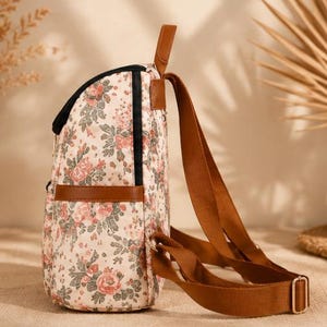 May include: A backpack featuring a cream-colored fabric with a floral pattern of pink roses and green leaves. The bag includes brown faux leather trim, a top handle, and adjustable brown webbing straps with gold-toned metal hardware and black zippers.