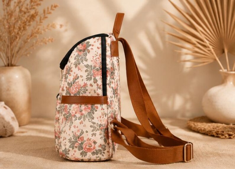 May include: A backpack featuring a cream-colored fabric with a floral pattern of pink roses and green leaves. The bag includes brown faux leather trim, a top handle, and adjustable brown webbing straps with gold-toned metal hardware and black zippers.