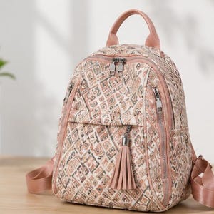 May include: A small backpack featuring a quilted texture with a geometric pattern in shades of beige, brown, and muted pink. It includes a top handle, adjustable shoulder straps, silver-tone metal zippers, and a decorative tassel on the front pocket.
