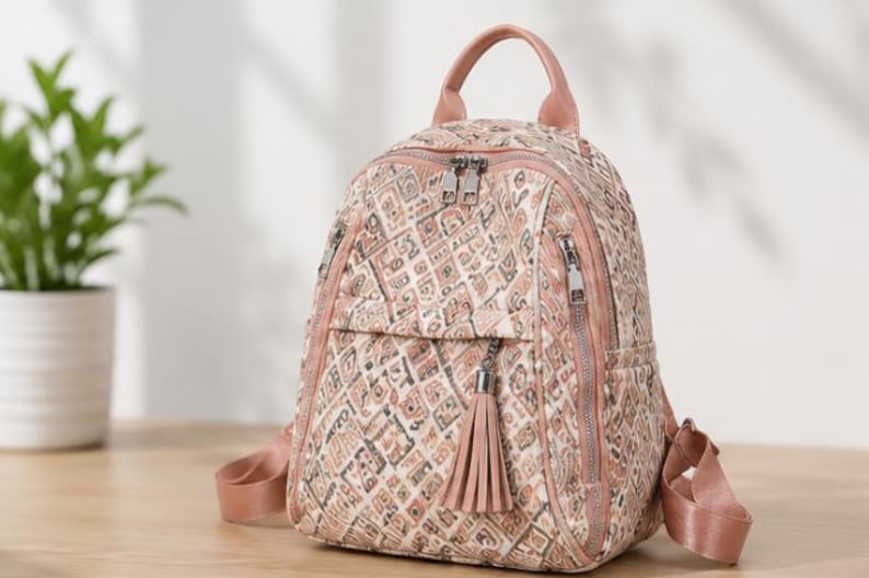 May include: A small backpack featuring a quilted texture with a geometric pattern in shades of beige, brown, and muted pink. It includes a top handle, adjustable shoulder straps, silver-tone metal zippers, and a decorative tassel on the front pocket.
