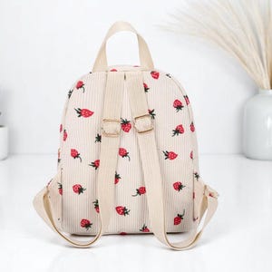 May include: A small beige corduroy backpack with adjustable straps. The backpack has a strawberry print all over it.