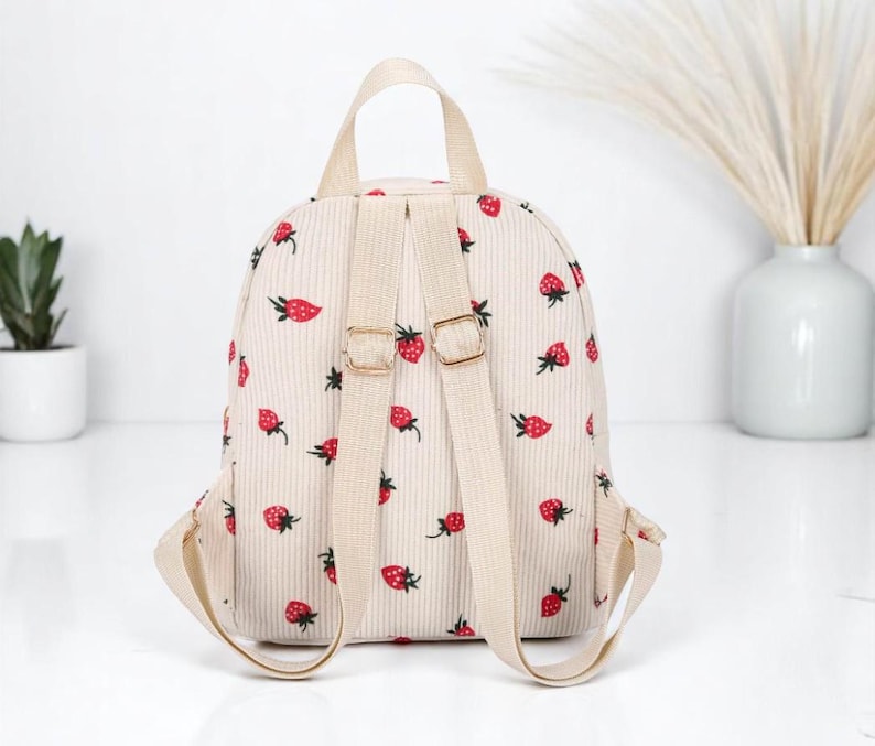 May include: A small beige corduroy backpack with adjustable straps. The backpack has a strawberry print all over it.