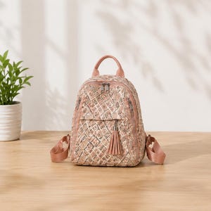 May include: A small backpack featuring a quilted texture with a repeating geometric pattern in shades of pink, beige, and brown. The bag includes a top handle, adjustable shoulder straps, silver-tone metal zippers, and a decorative tassel on the front pocket.