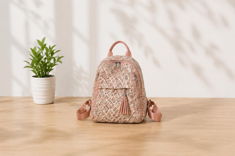 May include: A small backpack featuring a quilted texture with a repeating geometric pattern in shades of pink, beige, and brown. The bag includes a top handle, adjustable shoulder straps, silver-tone metal zippers, and a decorative tassel on the front pocket.